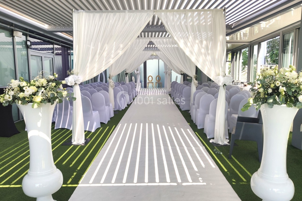 Allée centrale décorée pour une cérémonie, avec chaises alignées, rideaux blancs et arrangements floraux.
