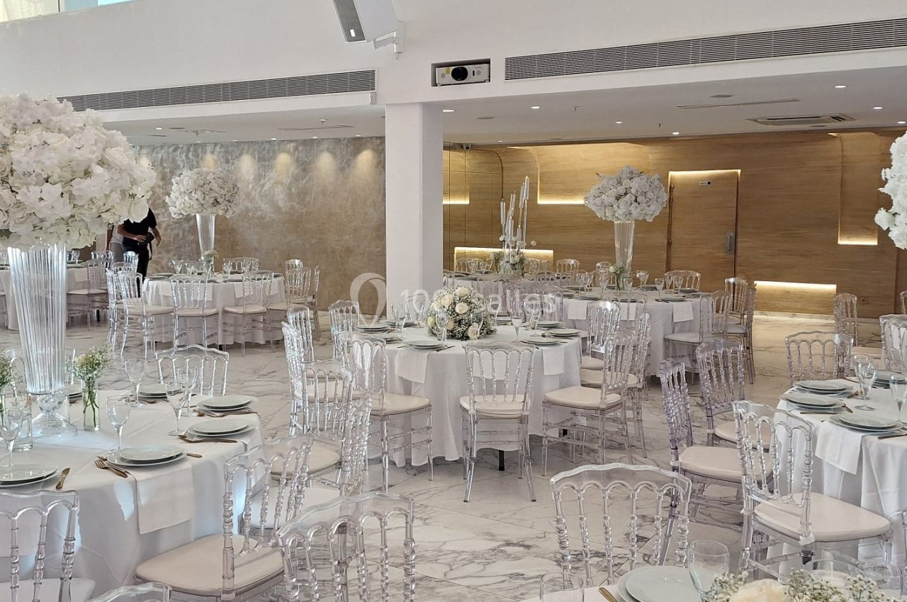 Salle de réception élégante avec tables rondes dressées, chaises transparentes et décorations florales blanches.
