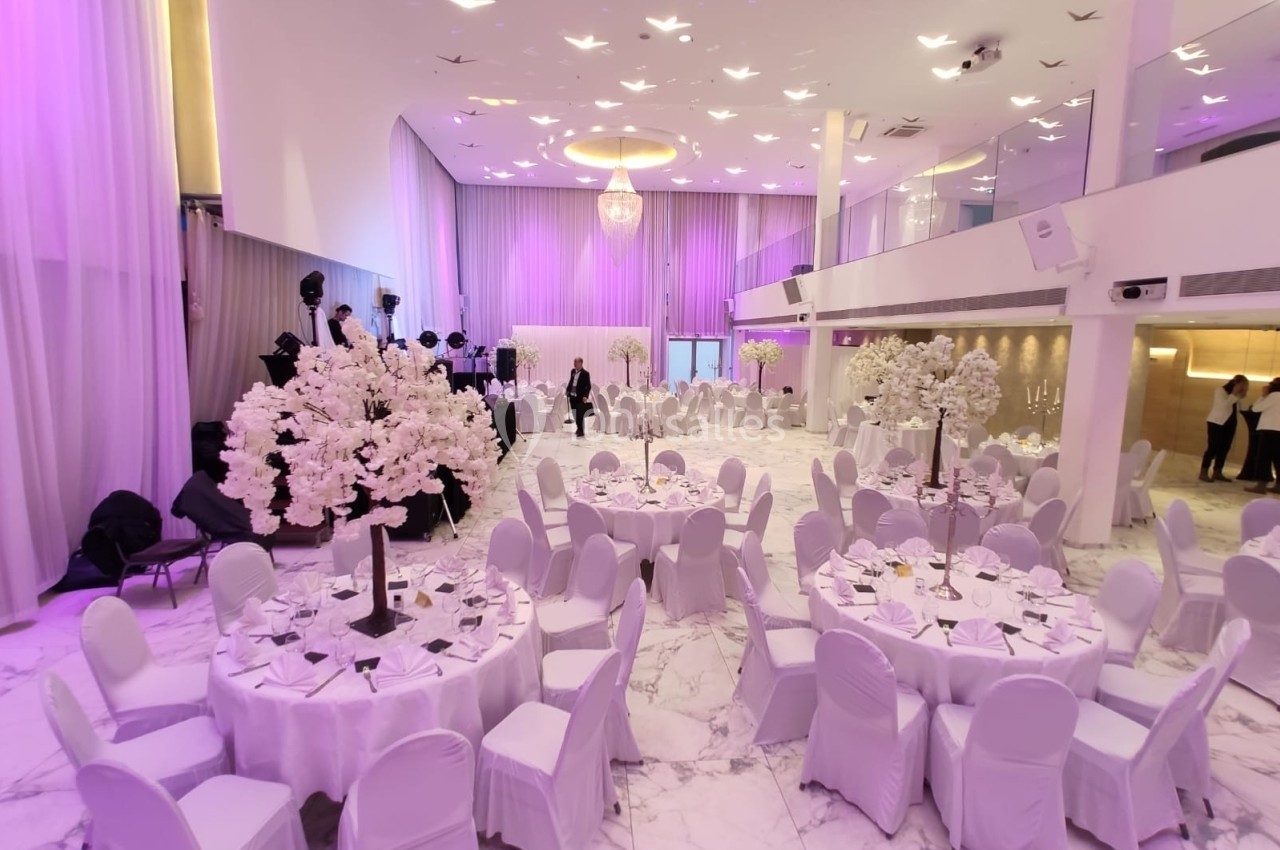 Salle de réception élégante avec tables rondes dressées, chaises blanches et décorations florales sous un éclairage violet.