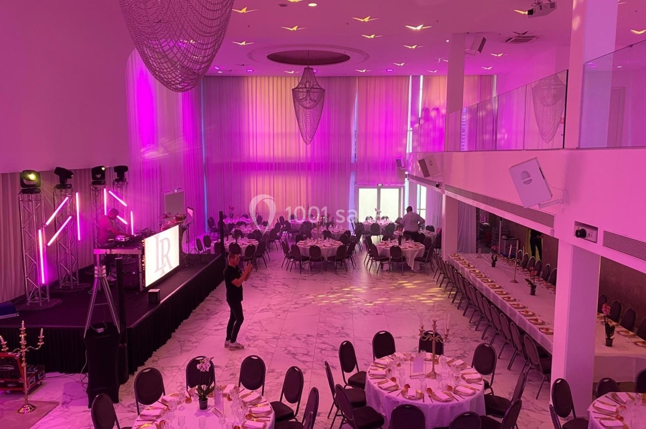 Salle de réception décorée avec des tables rondes dressées, éclairage rose et scène équipée pour un événement.