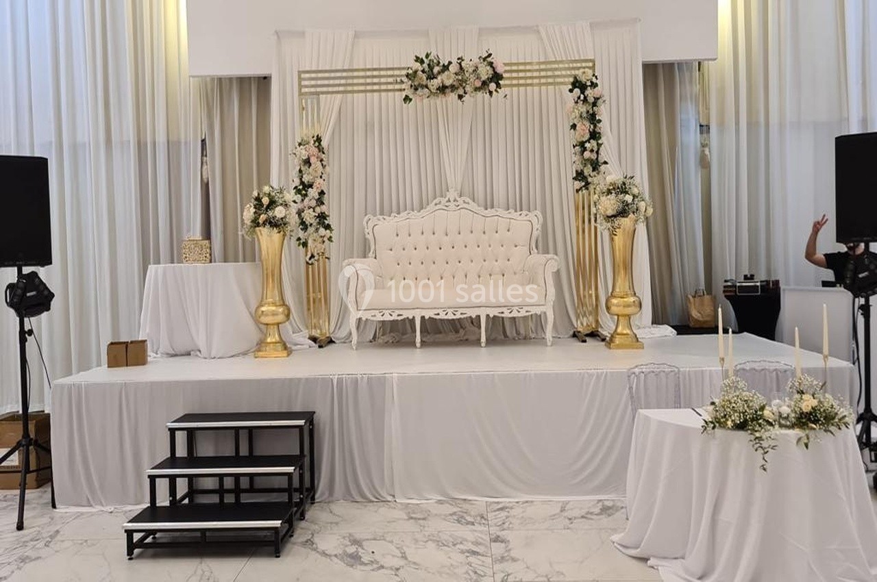 Estrade décorée pour un événement avec canapé blanc, colonnes dorées ornées de fleurs et rideaux blancs en arrière-plan.