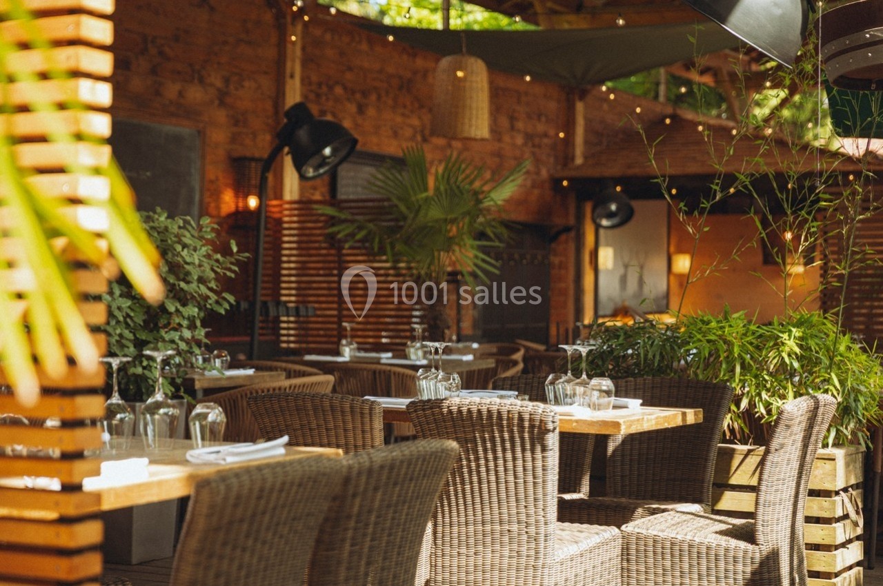 Terrasse de restaurant avec tables en bois, chaises en osier, plantes vertes et éclairage chaleureux.