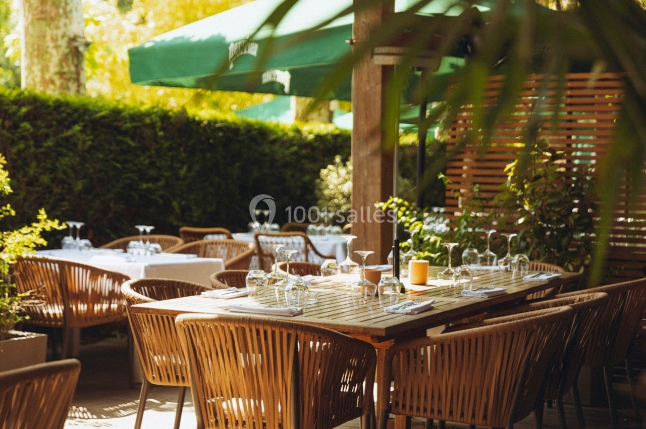 Terrasse d'un restaurant avec des tables en bois dressées, entourée de végétation et protégée par des parasols verts.