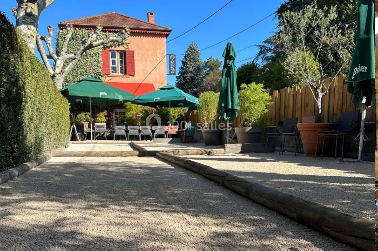 Terrasse extérieure avec terrain de pétanque, parasols verts, chaises et bâtiment rouge en arrière-plan.