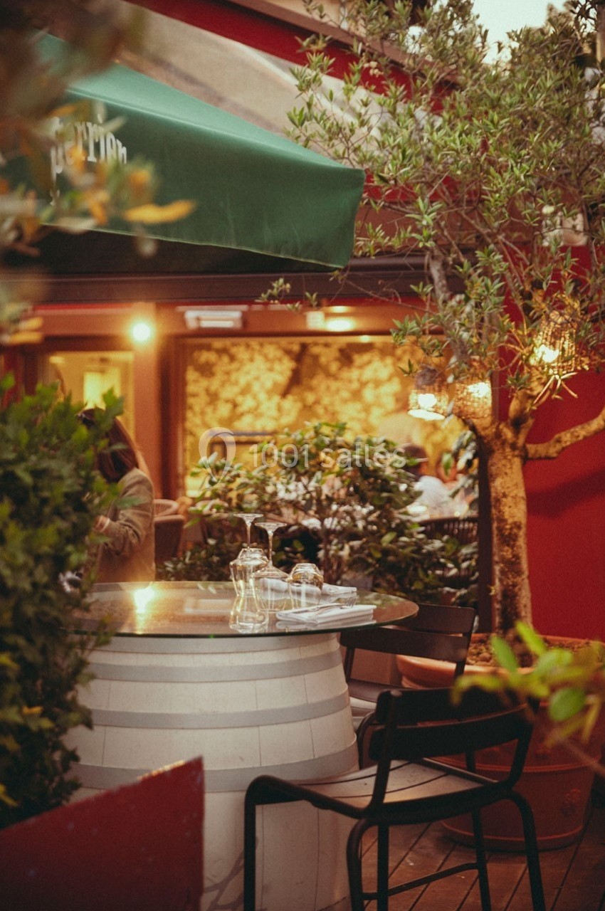 Terrasse de restaurant avec table ronde en verre sur un tonneau, chaises noires et végétation environnante.