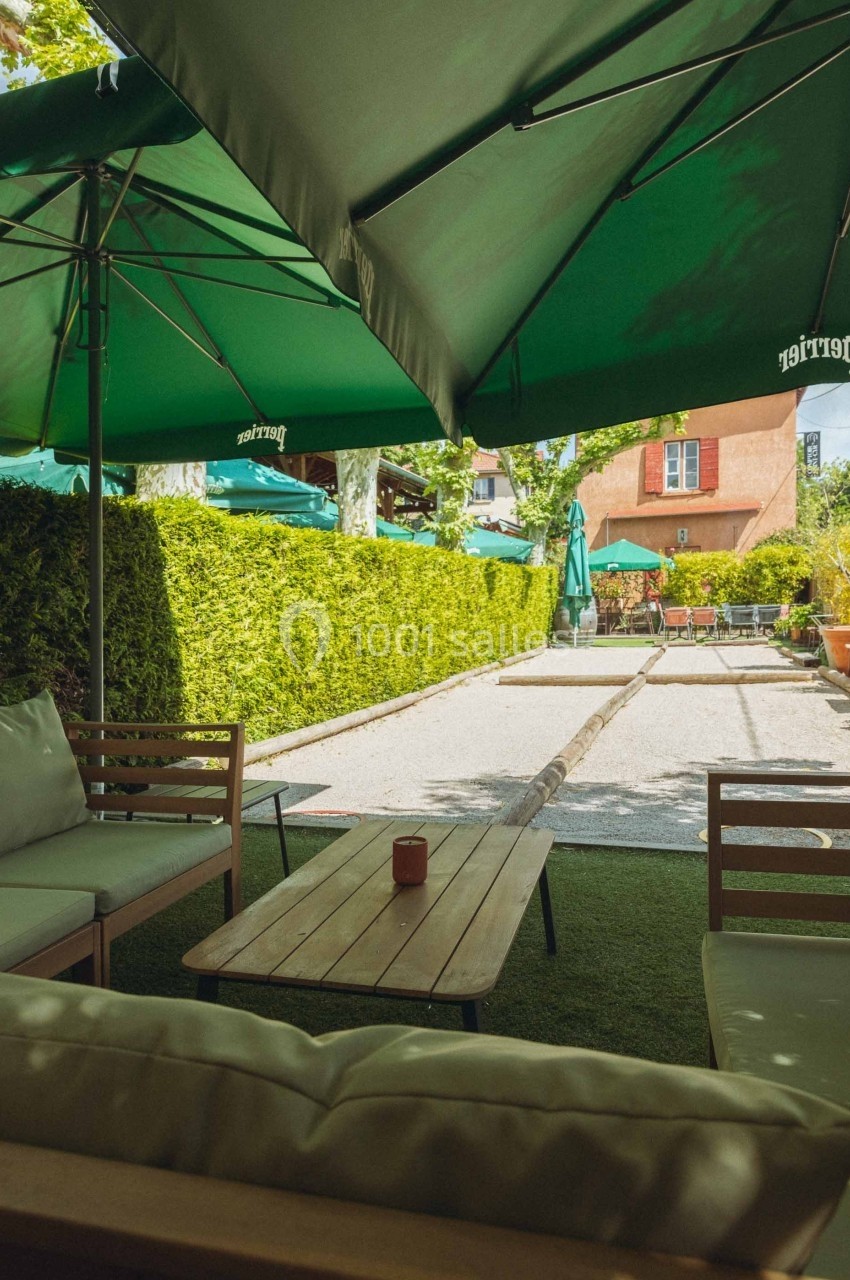 Terrasse extérieure avec tables en bois, chaises et parasols verts, entourée de haies et d'arbres.