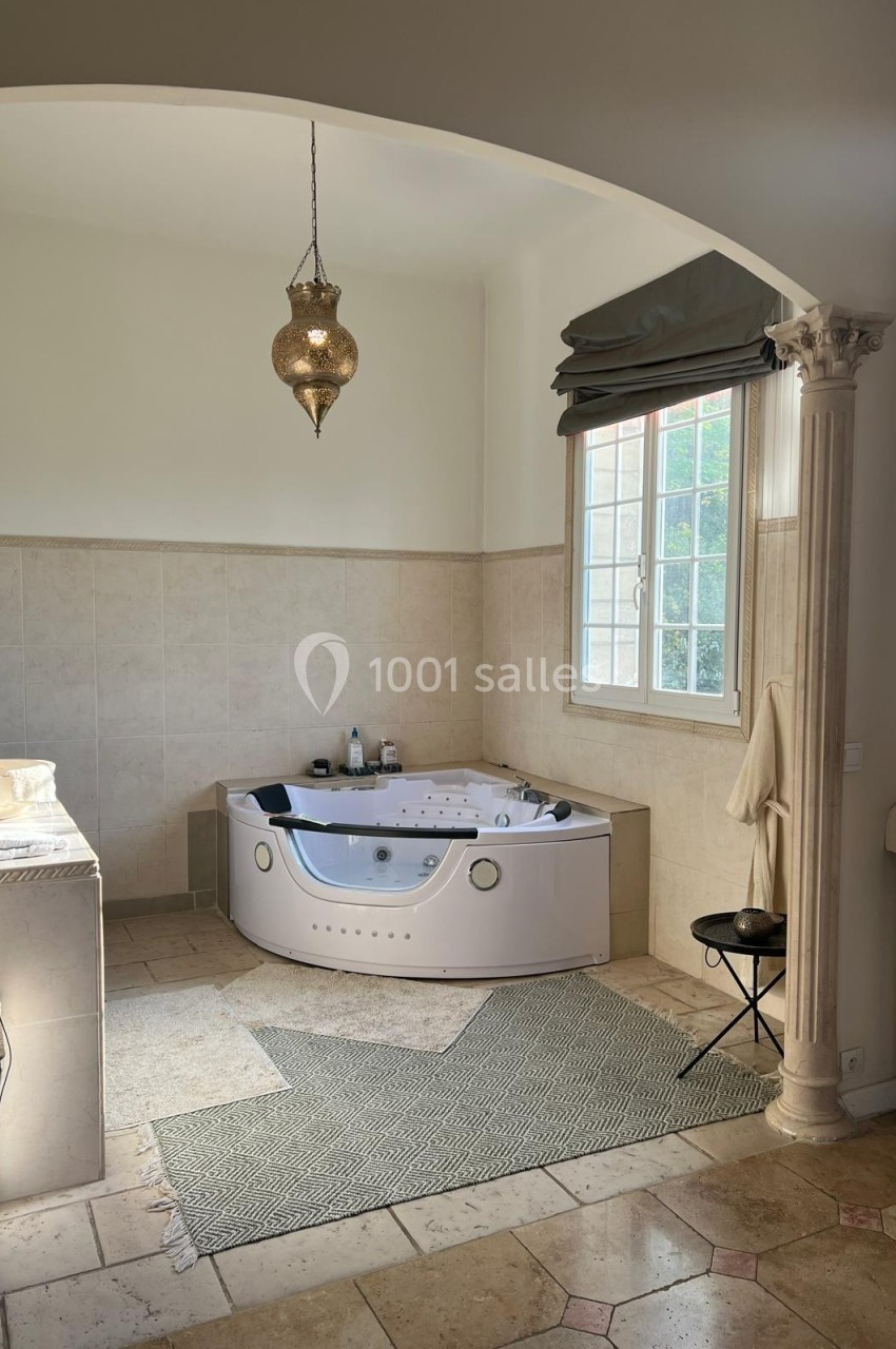 Salle de bain lumineuse avec baignoire balnéo, carrelage beige, fenêtre à rideaux et suspension décorative dorée.