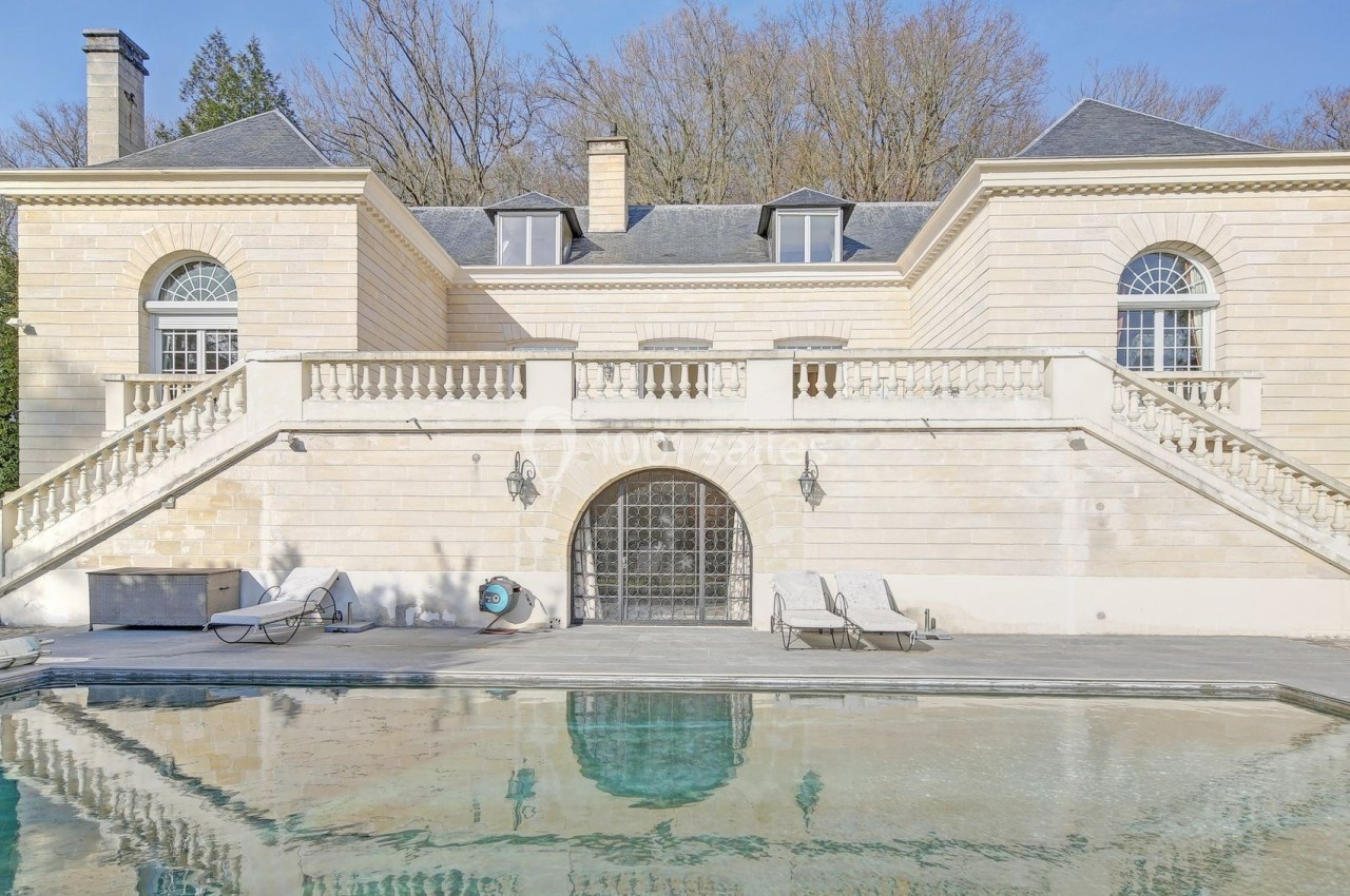 Façade d'une maison en pierre avec escalier symétrique, piscine au premier plan et arbres en arrière-plan.
