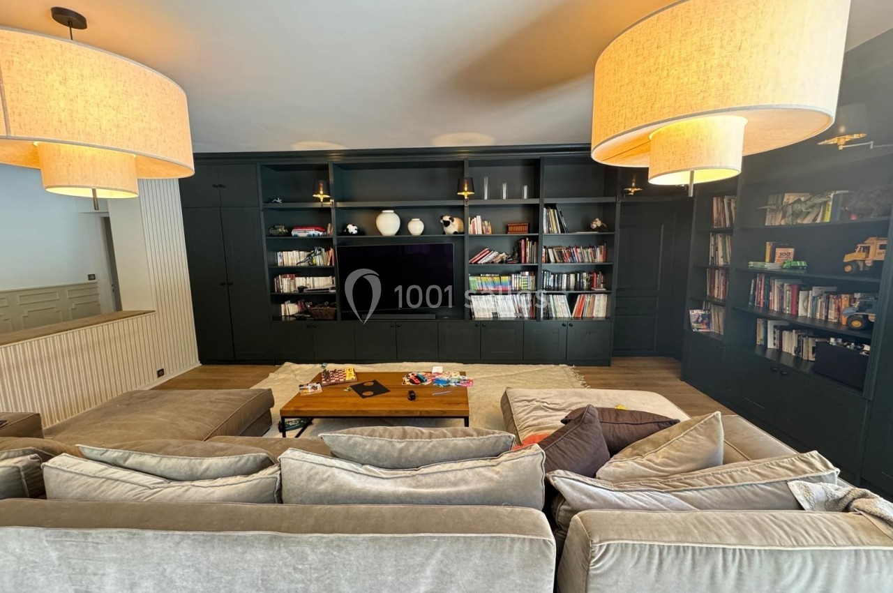 Salon avec canapé beige, table basse en bois, étagères noires remplies de livres et jouets, éclairé par des suspensions.
