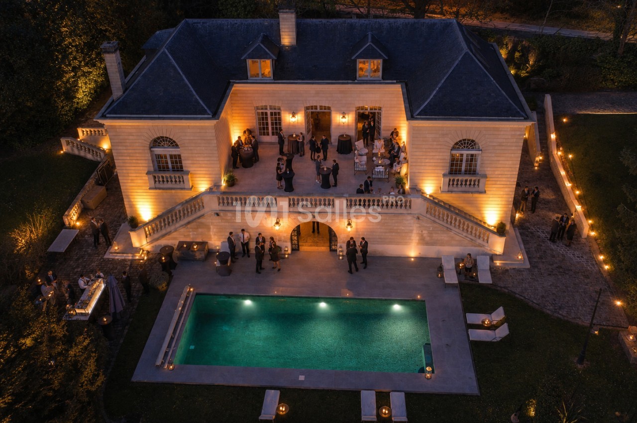 Vue aérienne d'une maison éclairée en soirée avec une piscine et des invités rassemblés sur la terrasse et dans le jardin.