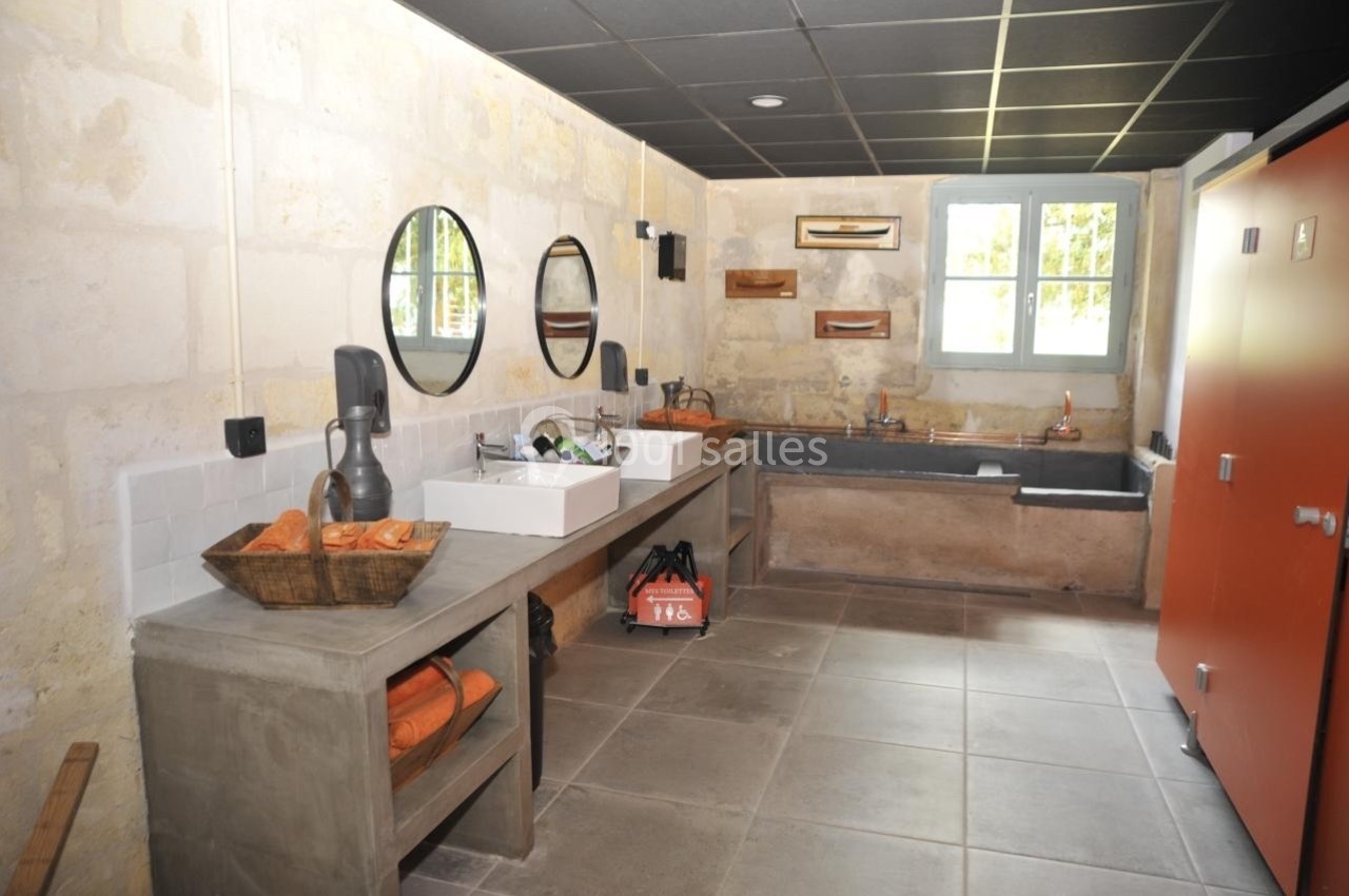 Salle de bain avec murs en pierre, deux lavabos, miroirs ronds, baignoire en béton et rangements avec serviettes orange.