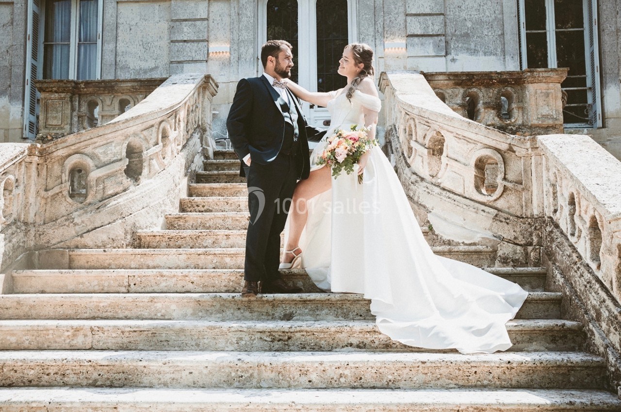 Un couple de mariés pose sur un escalier en pierre devant un bâtiment ancien, la mariée tenant un bouquet coloré.