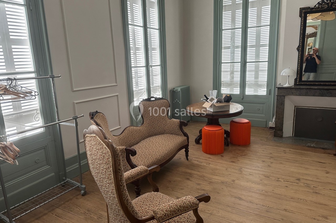 Salon lumineux avec parquet, mobilier ancien, table ronde, tabourets orange et miroir encadré sur un mur clair.