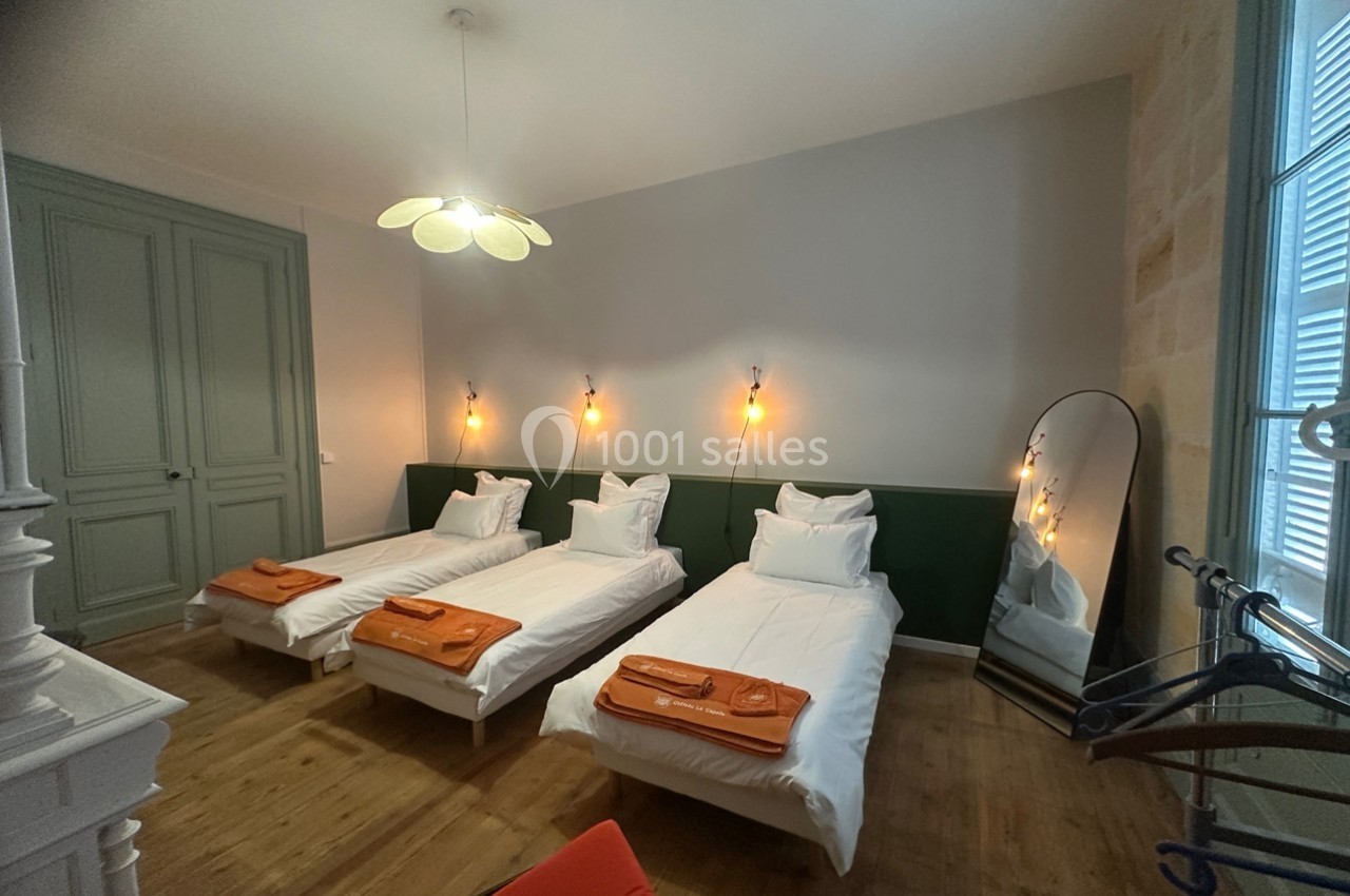 Chambre avec trois lits simples, linge orange plié, miroir sur pied, éclairage mural et parquet en bois.