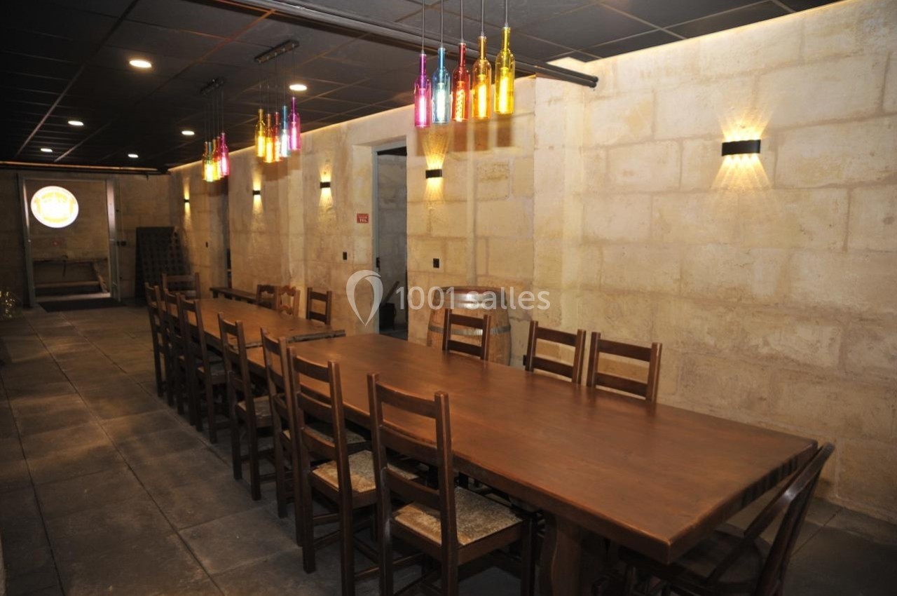 Salle avec une longue table en bois entourée de chaises, éclairée par des lampes murales et des bouteilles suspendues.