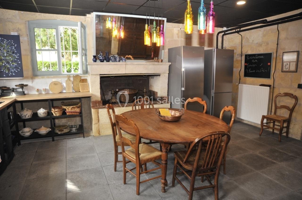 Cuisine rustique avec table en bois, cheminée ancienne, étagères ouvertes, réfrigérateurs modernes et suspensions colorées.