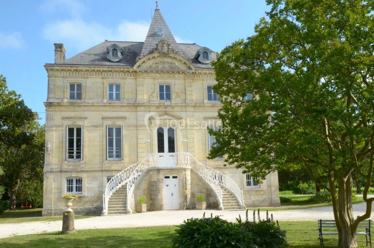Façade d'un manoir en pierre claire avec escalier central, entouré d'arbres et d'un jardin verdoyant.