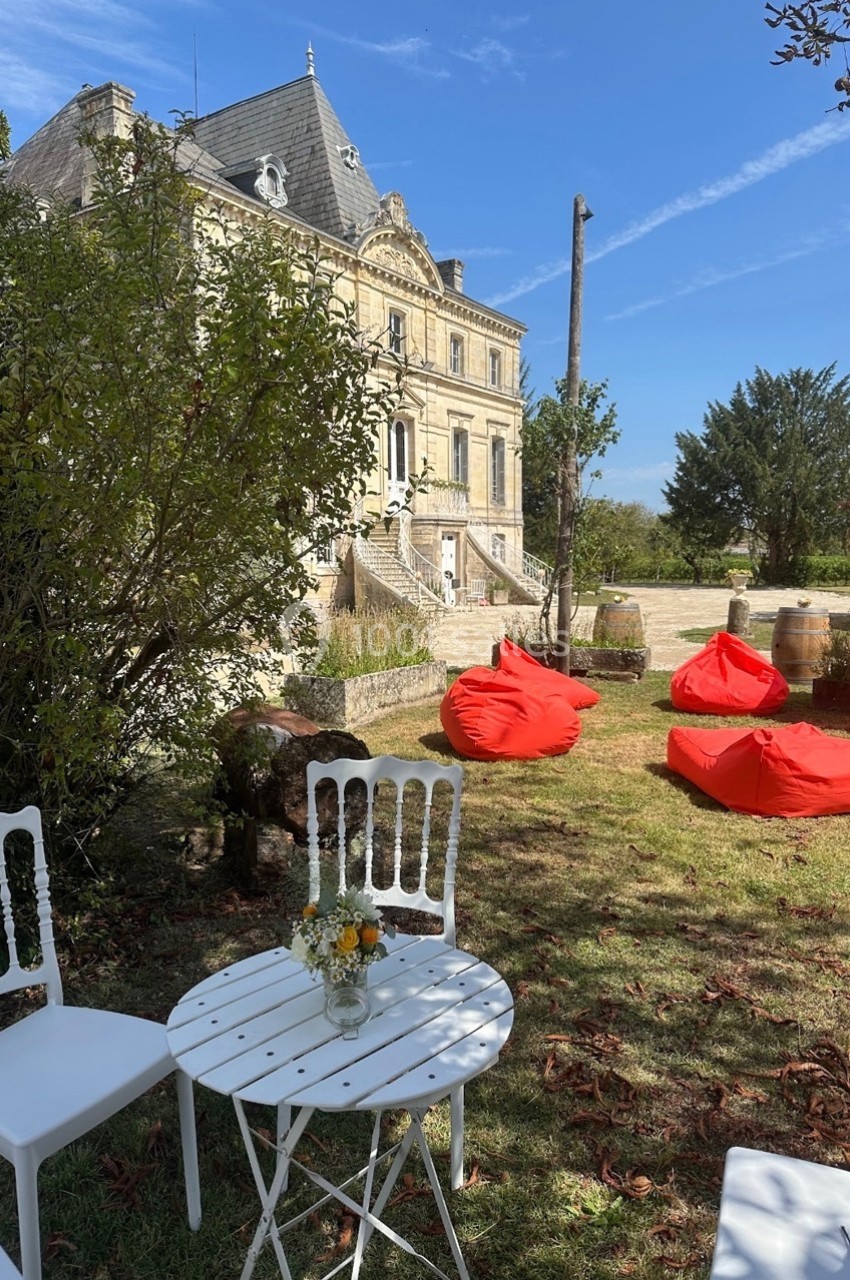 Chaises blanches et table ronde dans un jardin, avec des poufs rouges et un bâtiment ancien en arrière-plan.