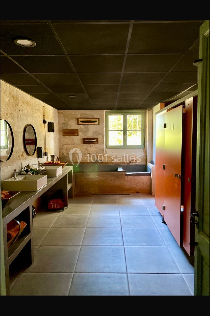 Salle de bain lumineuse avec murs en pierre, lavabos modernes, cabines orange et fenêtre laissant entrer la lumière…