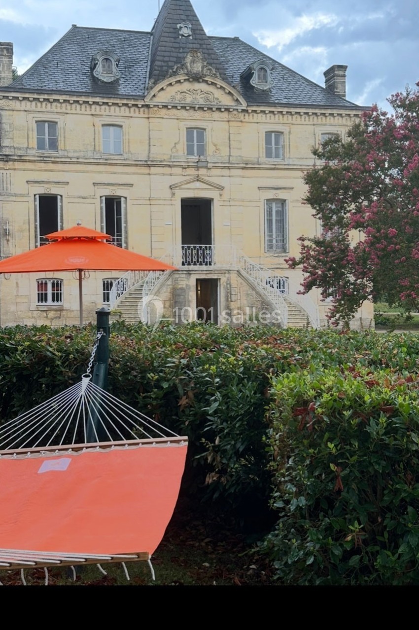 Hamac orange installé dans un jardin devant une grande maison en pierre avec un toit mansardé et des arbres fleuris.