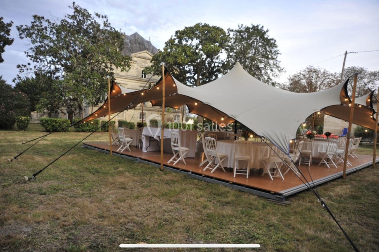 Tente élégante installée sur une plateforme en bois, abritant des tables et chaises dans un jardin verdoyant.