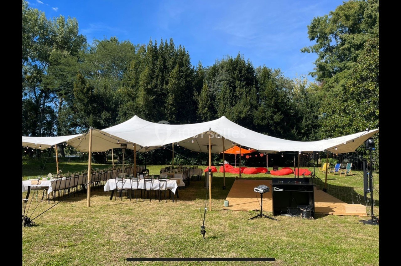 Grande tente blanche installée dans un jardin, abritant des tables dressées et un espace avec des coussins rouges.