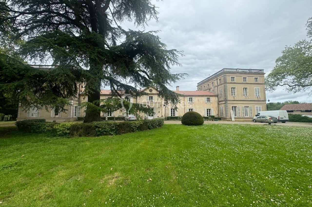Façade d'un grand bâtiment ancien entouré d'une pelouse verte et d'arbres sous un ciel nuageux.