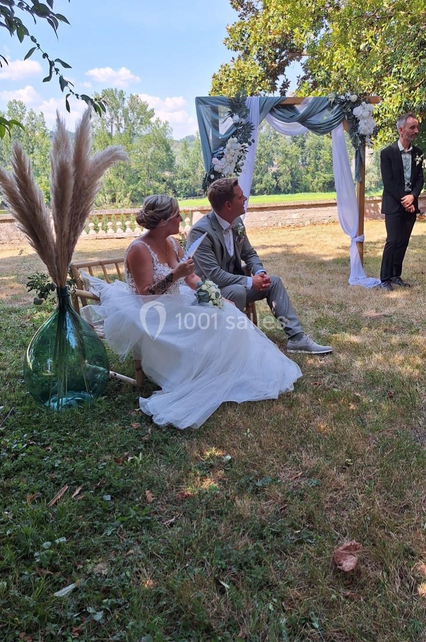 Un couple assis en tenue de mariage sous une arche décorée, dans un jardin verdoyant par une journée ensoleillée.