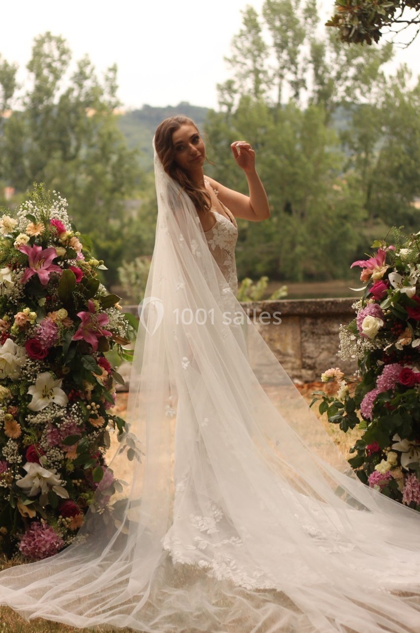 Femme en robe de mariée avec un long voile, debout entre deux compositions florales colorées en extérieur.