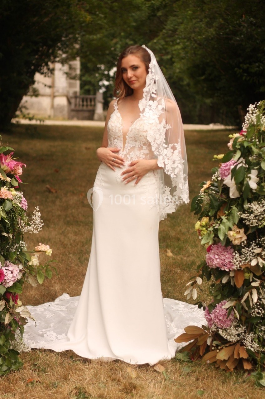Femme en robe de mariée blanche avec voile en dentelle, debout dans un jardin entouré de compositions florales.