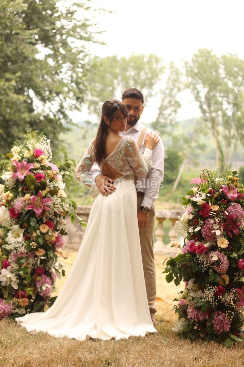 Un couple pose entre deux compositions florales colorées dans un cadre extérieur verdoyant.
