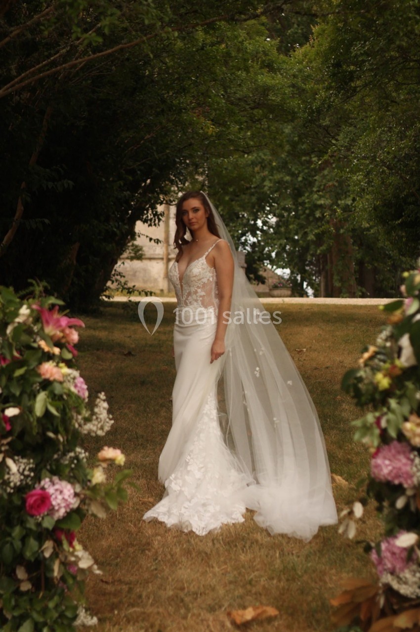 Femme en robe de mariée blanche avec un voile, debout dans un jardin entouré de fleurs et d'arbres.