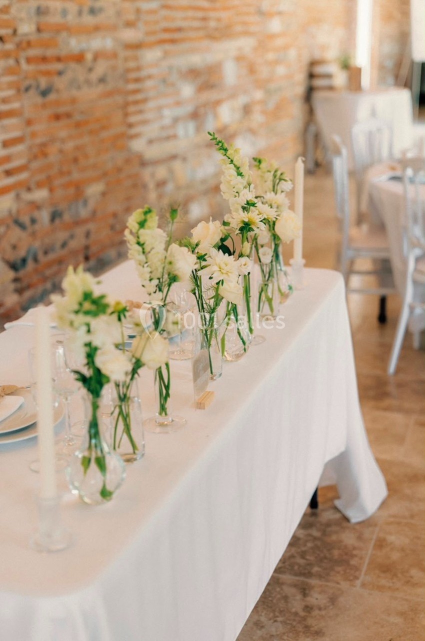 Table décorée avec des vases de fleurs blanches et bougies, dans une salle aux murs en briques apparentes.