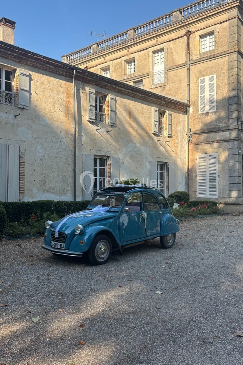 Voiture ancienne bleue garée devant une grande maison en pierre avec volets blancs, sous un ciel dégagé.