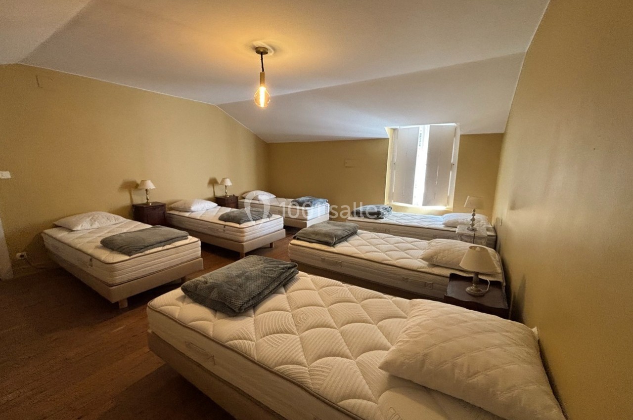 Dortoir avec six lits simples alignés, draps et couvertures gris, éclairage naturel et lampe suspendue.