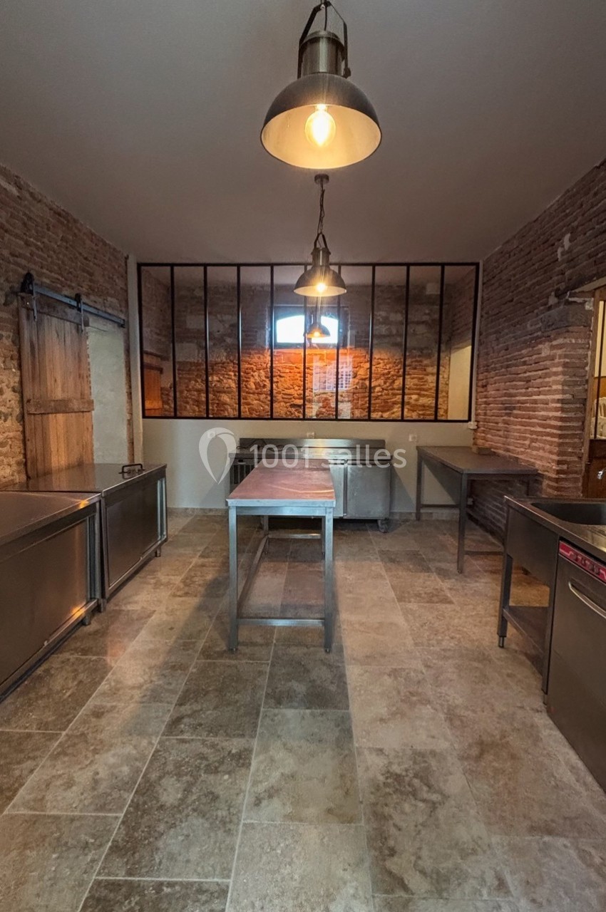 Cuisine professionnelle avec murs en briques, plan de travail en métal, sol en carrelage et éclairage suspendu.