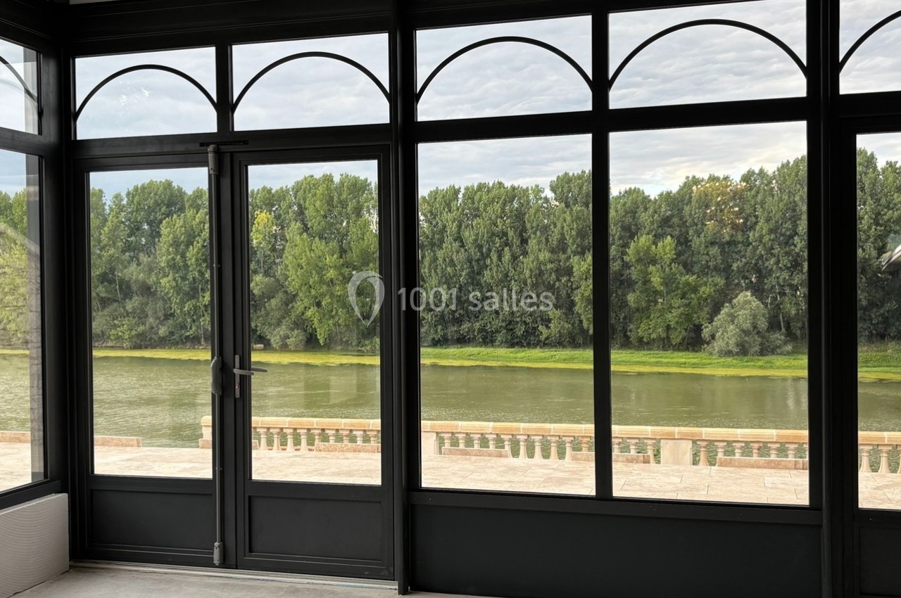 Vue d'une rivière bordée d'arbres, observée depuis une véranda aux grandes baies vitrées.