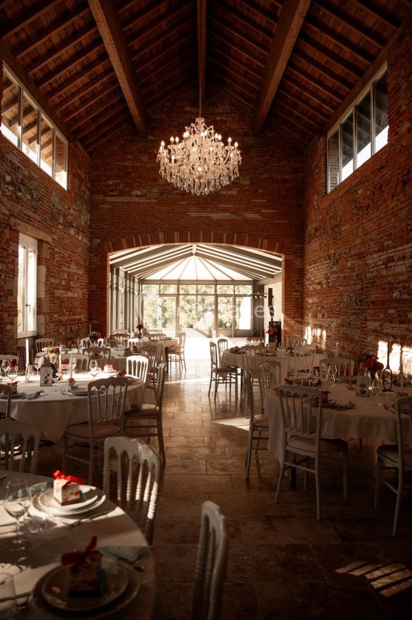 Salle de réception en briques avec tables dressées, chaises blanches, lustre central et lumière naturelle entrant par une…