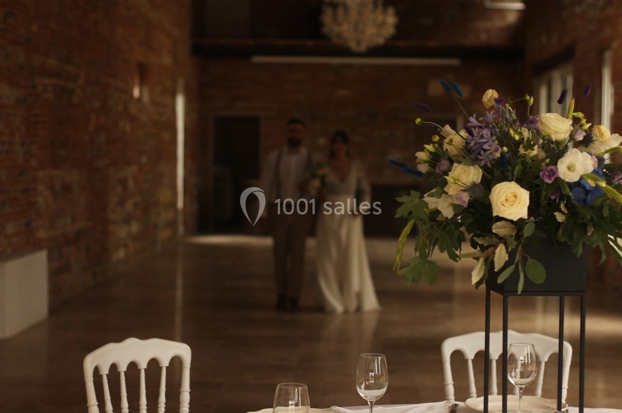 Un couple en tenue de mariage marche dans une salle en briques, avec un bouquet de fleurs au premier plan.