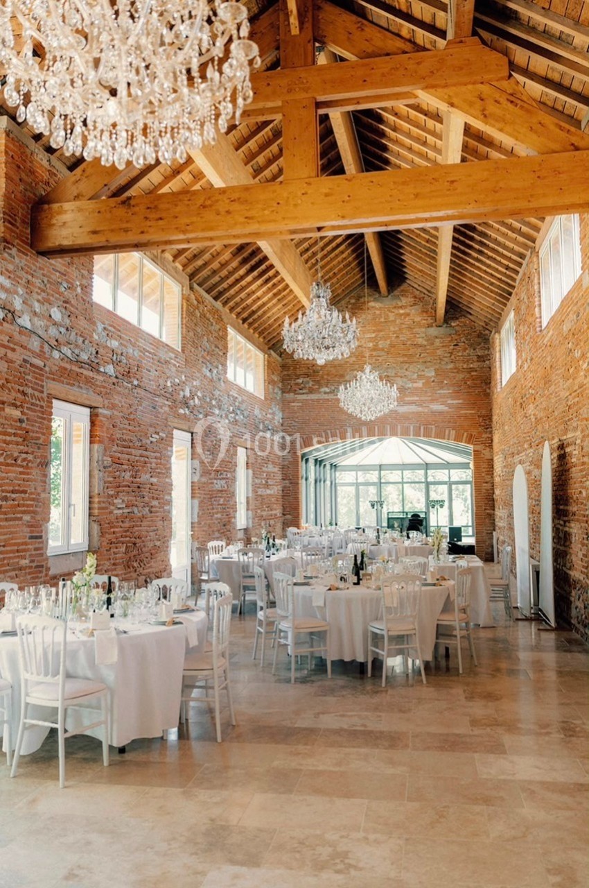 Salle de réception lumineuse avec murs en briques, poutres apparentes, lustres et tables dressées pour un événement.