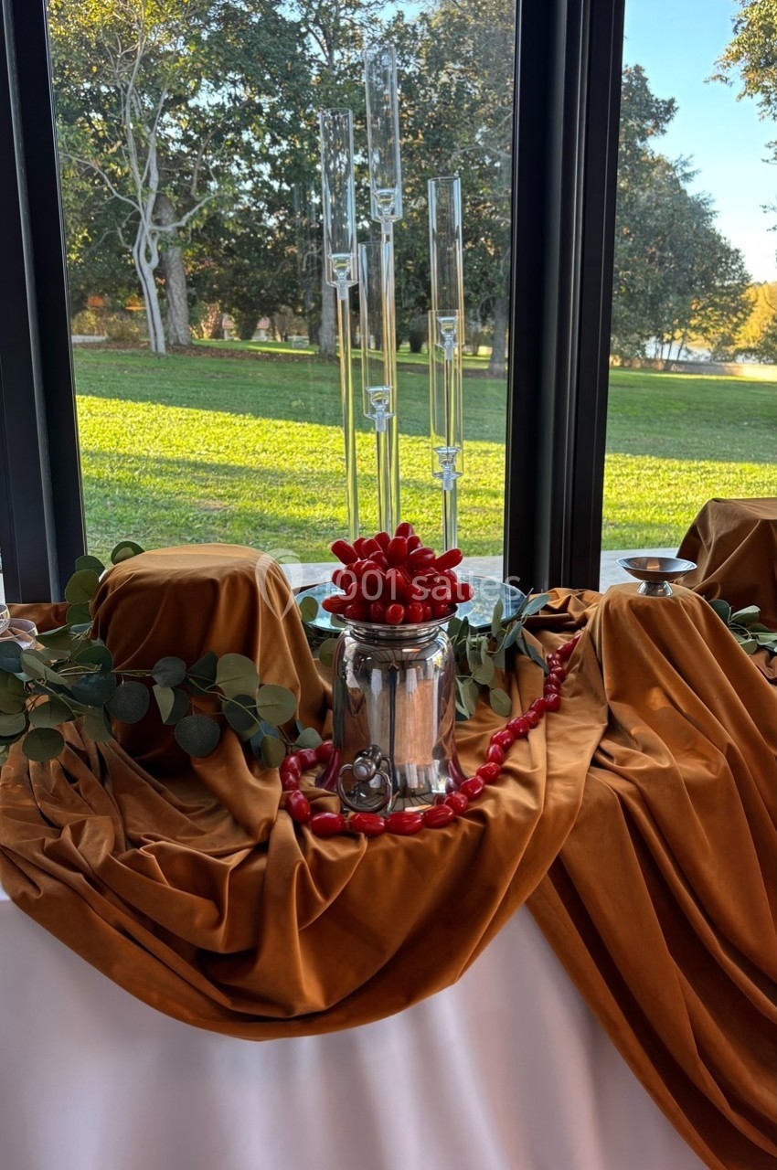 Table décorée avec des drapés ocres, un vase argenté rempli de cerises rouges et des bougies en verre, devant une fenêtre…
