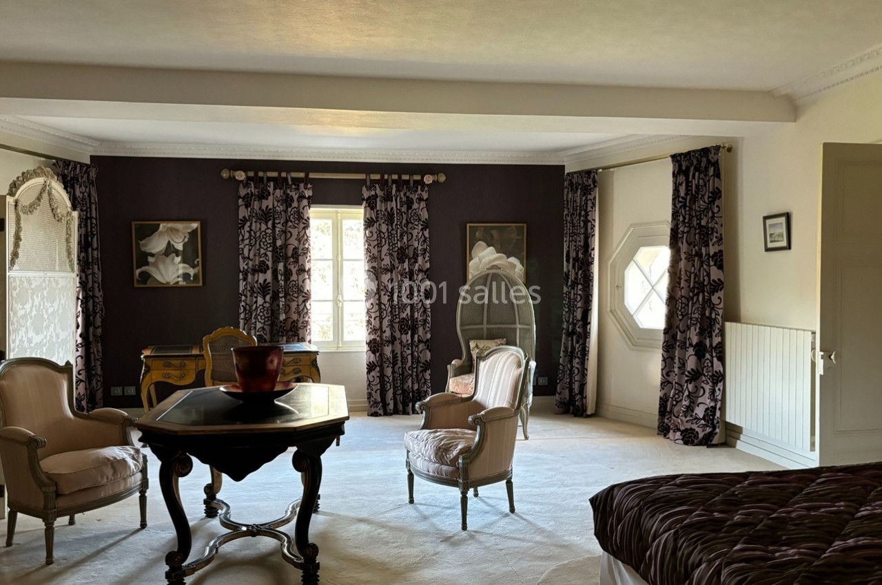 Chambre spacieuse avec mobilier classique, rideaux à motifs sombres, bureau ancien et fauteuils autour d'une table.