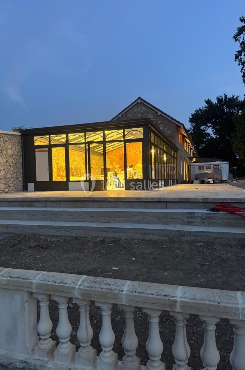 Extension vitrée d'un bâtiment en pierre, éclairée de l'intérieur au crépuscule, avec un espace extérieur en travaux.