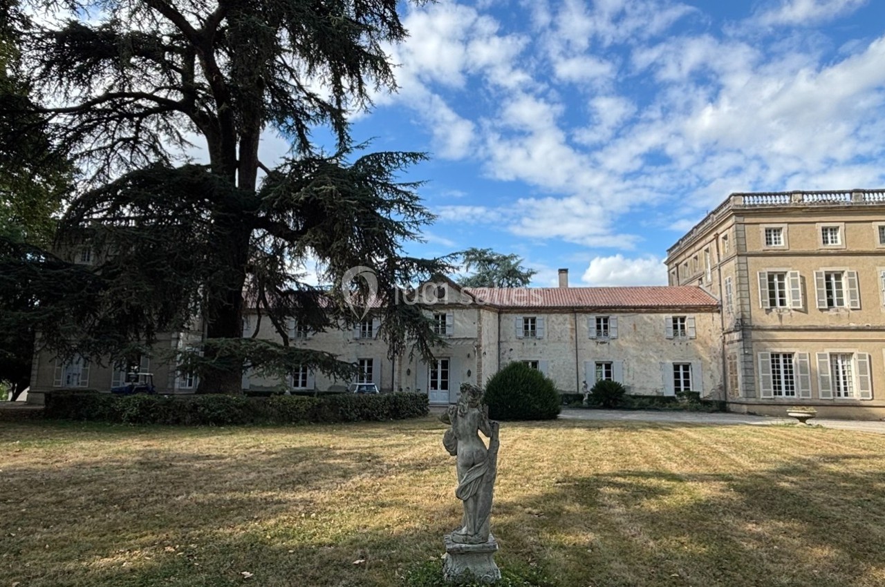 Façade d'un grand bâtiment en pierre entouré d'arbres, avec une statue au premier plan sur une pelouse.