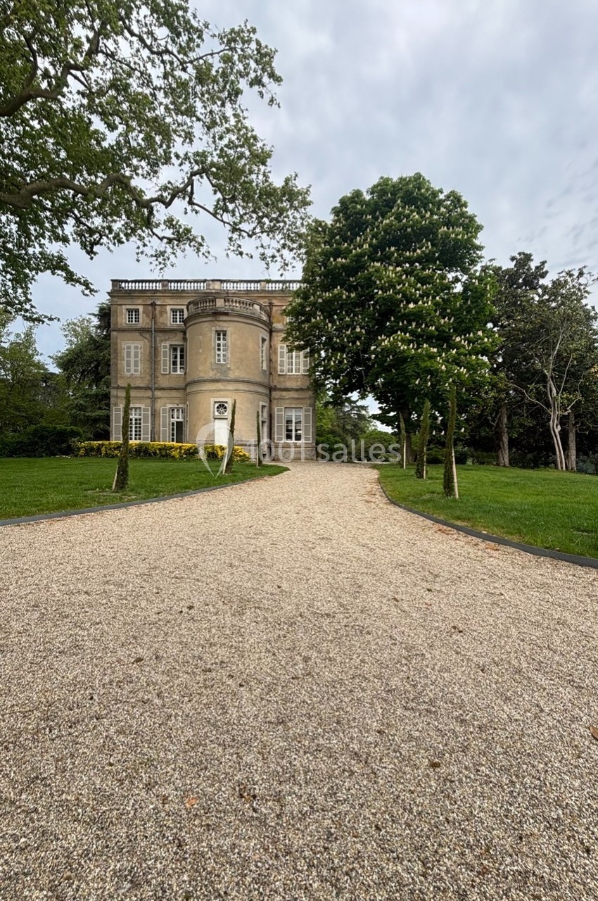 Allée gravillonnée menant à une maison ancienne entourée d'arbres et de pelouses sous un ciel nuageux.