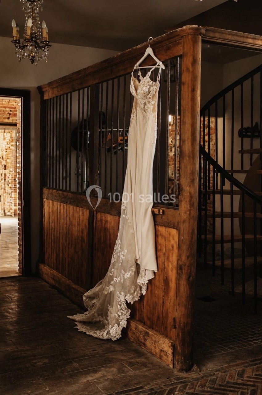 Robe de mariée en dentelle suspendue à une porte en bois dans un intérieur rustique avec escalier en colimaçon.