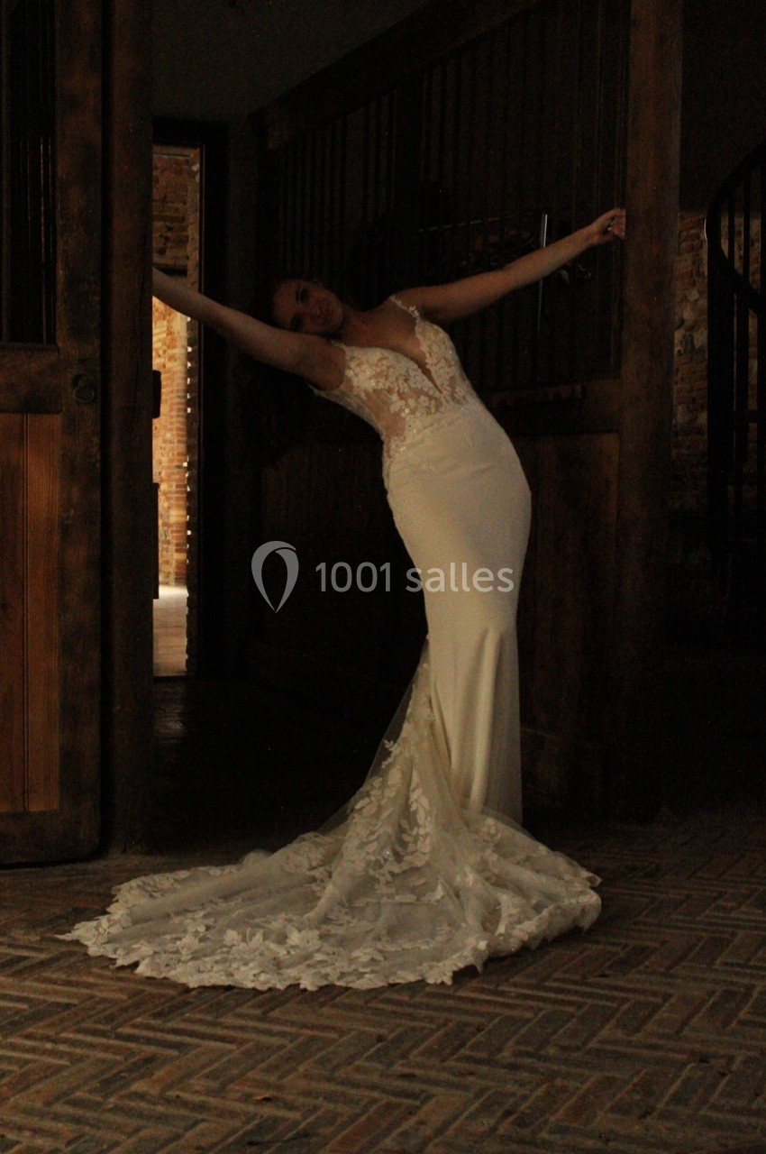Femme en robe de mariée ajustée avec une longue traîne en dentelle, posant dans un intérieur sombre en brique.