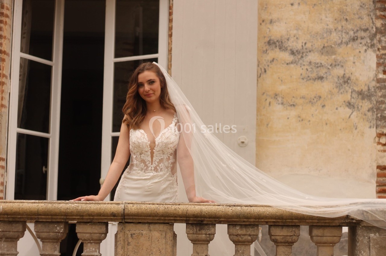 Femme en robe de mariée blanche avec un voile long, posant devant une balustrade en pierre et un mur texturé.