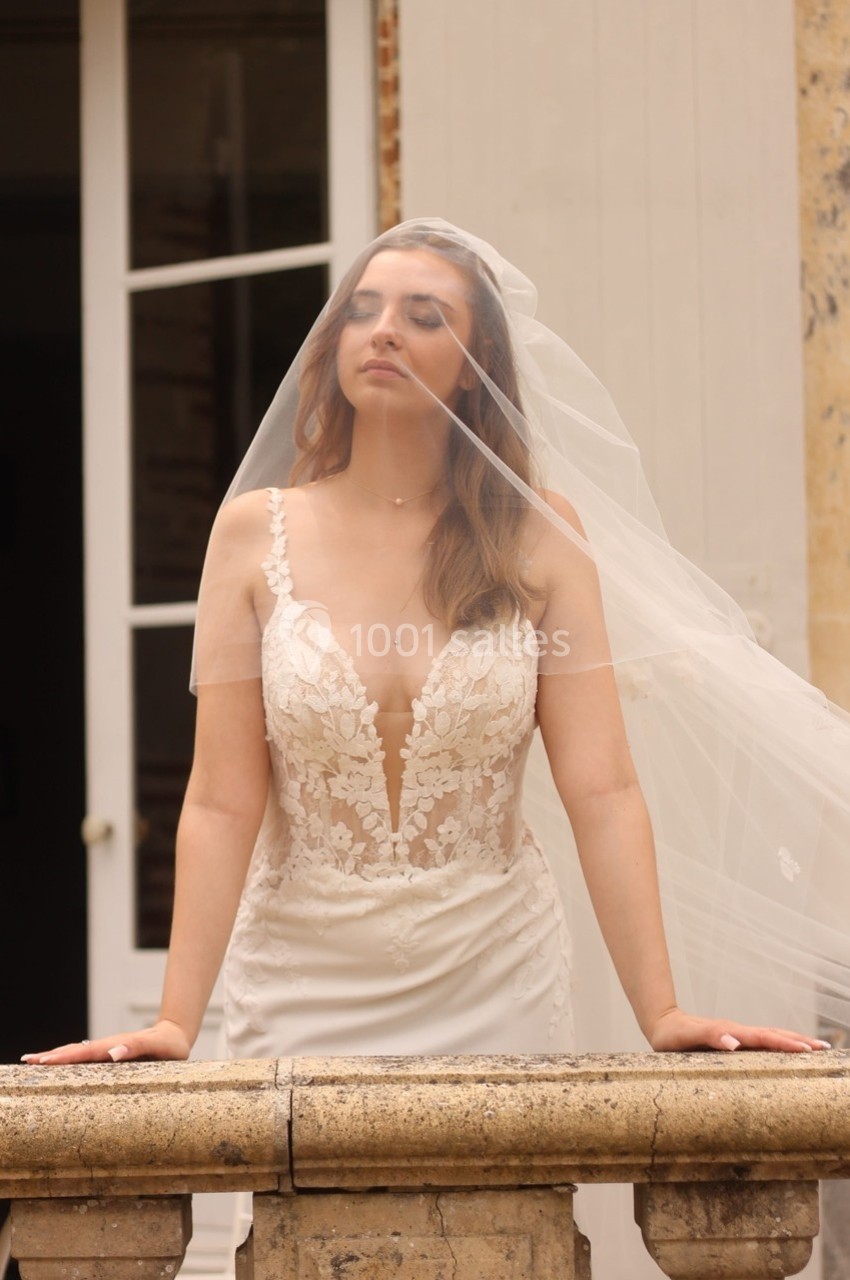 Femme en robe de mariée avec un voile, debout devant une balustrade en pierre, les yeux fermés.