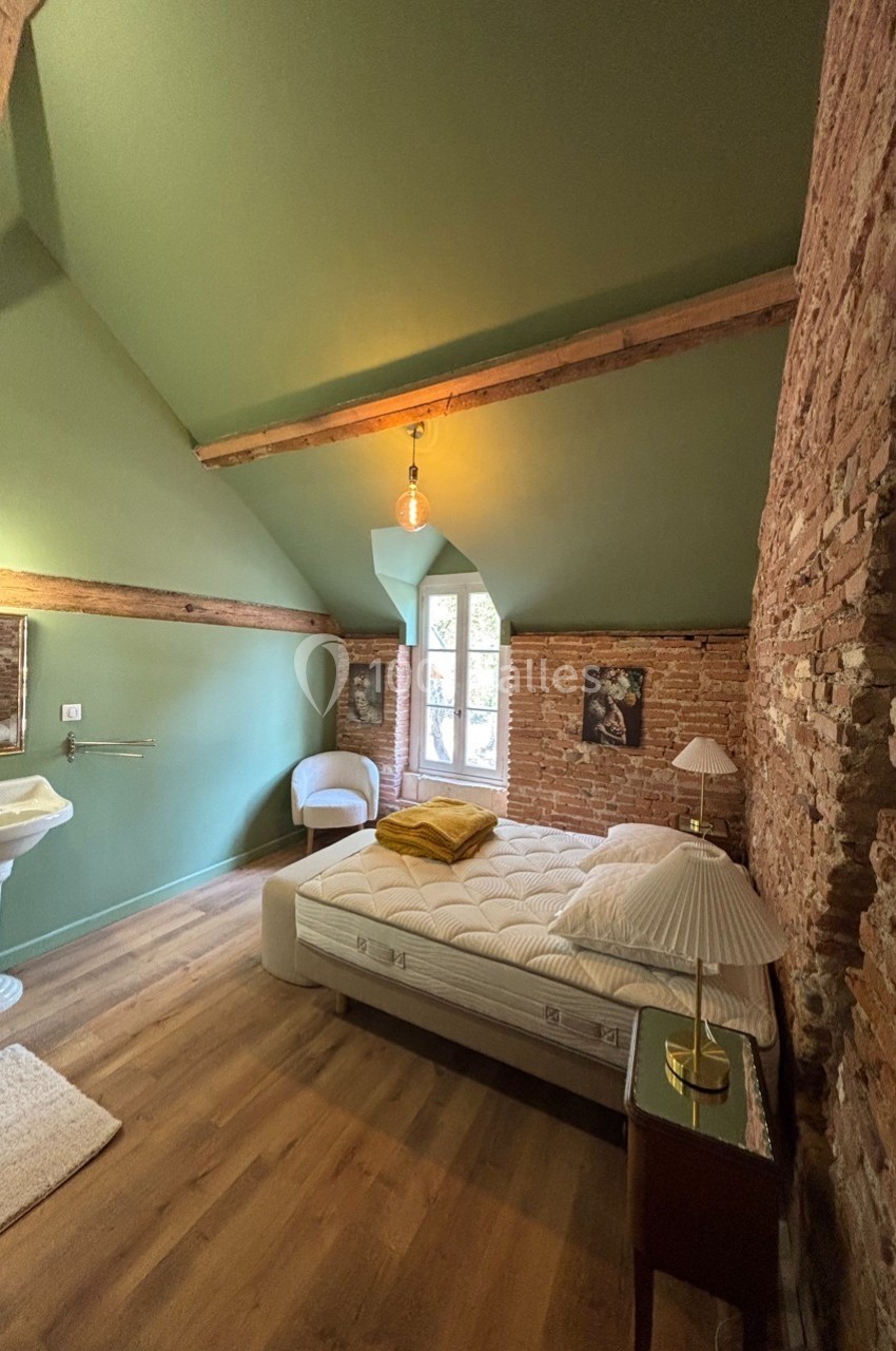 Chambre avec murs en briques, plafond vert, lit double, lampes de chevet et grande fenêtre laissant entrer la lumière.